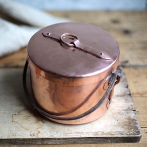 May include: A copper pot with a lid and a handle. The pot is round and has a shiny finish. The lid has a handle and a loop. The pot is sitting on a wooden surface.