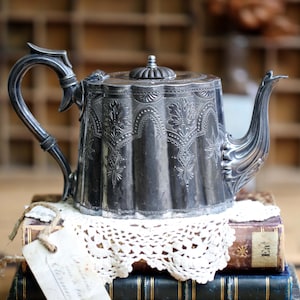 Könnte beinhalten: Eine silberne Teekanne mit kunstvollen floralen Details steht auf einem Stapel antiker Bücher und einer weißen Spitzendecke. Ein Anhänger hängt an den Büchern. Die Teekanne hat einen gebogenen Griff und Ausguss.