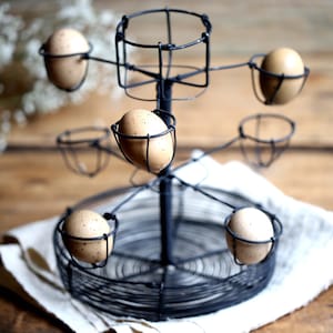May include: A black metal egg holder with six speckled brown eggs. The holder has a circular base and a Ferris wheel-like design with individual wire baskets for each egg. The holder sits on a white cloth, with a wooden background.