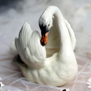 May include: A white ceramic swan figurine with a curved neck and outstretched wings. The swan's beak is orange and black, and the head is slightly tilted. The figurine is on a white surface with small white flowers.