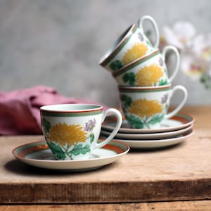 May include: A set of four white teacups and saucers. Each cup and saucer features a yellow chrysanthemum design with green leaves and a red and green border. The cups are stacked on top of each other.