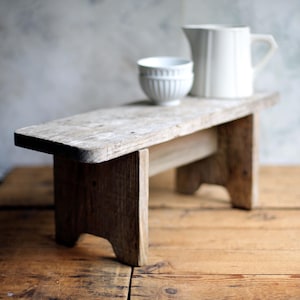 Puede incluir: Un taburete de madera rústico y desgastado con una parte superior rectangular y patas resistentes. Una jarra de cerámica blanca y un cuenco pequeño están sobre el taburete. El taburete es de madera marrón claro y está sobre una superficie de madera.