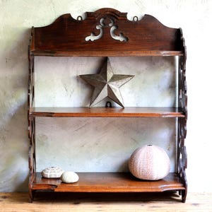 Könnte beinhalten: Ein dreistufiges Wandregal aus dunklem Holz mit dekorativen Ausschnitten und einer gewellten Oberseite. Die Regale beherbergen einen Metallstern, zwei kleine weiße Muscheln und einen runden, strukturierten Seeigel. Das Regal steht vor einer Wand in neutralen Tönen.