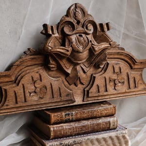 May include: A brown wooden architectural pediment featuring intricate carved floral and scroll motifs, resting atop a stack of three antique leather-bound books with gold-leaf detailing and a single light-colored feather placed at the base.