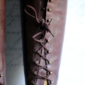 May include: A close-up of a brown leather boot with a lace-up design. The boot has a vintage look, with brass-colored eyelets and a dark brown lace. The boot is displayed on a wooden form.