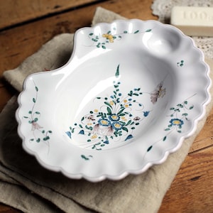 Op de afbeelding: Een witte, schelpvormige keramische schaal met een geschulpte rand. De schaal is versierd met een bloemmotief in blauw, geel en groen. Er is ook een vlinder afgebeeld. De schaal ligt op een beige linnen doek.