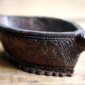 Peut inclure: Un bol en bois sculpté à la main, de couleur marron foncé, avec un bec et une poignée. Le bol présente des motifs sculptés complexes sur le bord et les côtés. Le bol en bois est présenté sur une surface en bois brun clair.