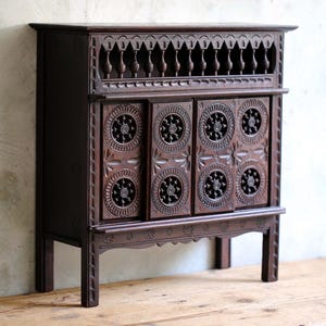Può includere: Un mobile in legno marrone scuro con quattro ante caratterizzate da intricati motivi circolari intagliati. La parte superiore include una fila di piccoli fusi torniti e la base inferiore è decorata con motivi floreali intagliati su quattro gambe dritte.