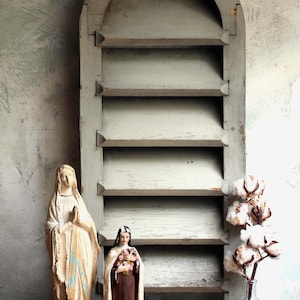 Könnte beinhalten: Ein rustikales, verwittertes Holzregal mit einem gewölbten Oberteil und mehreren Regalböden. Zwei religiöse Statuen stehen vor dem Regal, und eine kleine Glasflasche mit Baumwollstielen befindet sich auf der rechten Seite. Die Statuen sind in gedämpften Tönen bemalt.