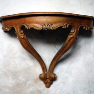 Puede incluir: Estantería de pared de madera marrón de estilo vintage, con patas curvas ornamentadas y un adorno floral decorativo en la parte inferior. La estantería tiene un borde frontal curvado. La madera tiene un aspecto cálido y envejecido. Ancho: aprox. 30 cm.