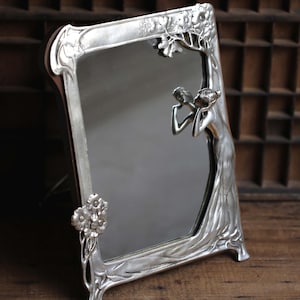 May include: A silver-toned, ornate mirror with a decorative frame featuring a stylized female figure and floral motifs. The mirror is freestanding and has a rectangular shape.