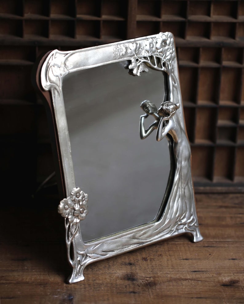 May include: A silver-toned, ornate mirror with a decorative frame featuring a stylized female figure and floral motifs. The mirror is freestanding and has a rectangular shape.
