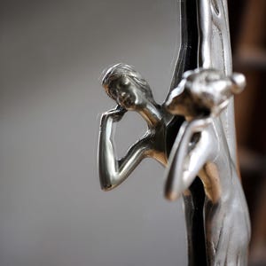 May include: A silver-toned mirror with a decorative frame featuring two figures of women sculpted in a tree. The mirror is oval shaped and has a reflective surface.