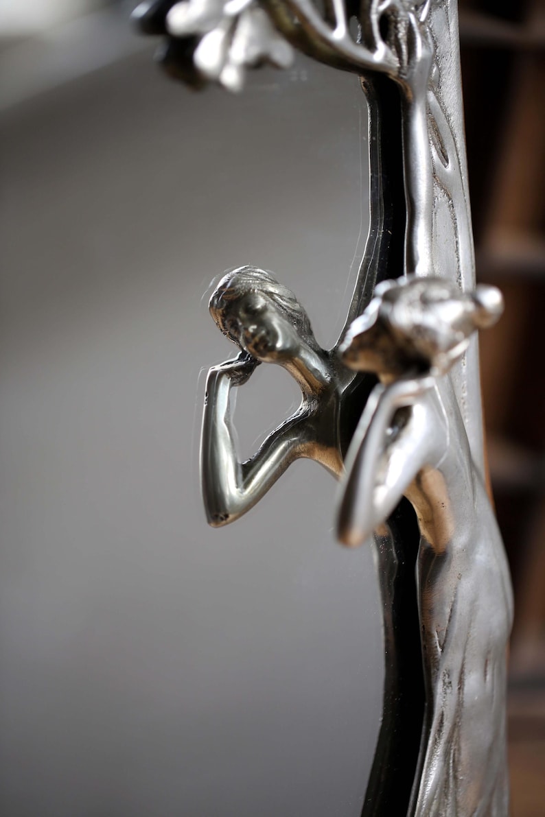 May include: A silver-toned mirror with a decorative frame featuring two figures of women sculpted in a tree. The mirror is oval shaped and has a reflective surface.