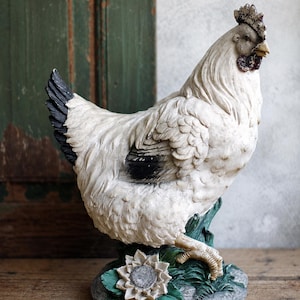 May include: A decorative figurine of a white chicken with black-tipped tail and wing feathers. The chicken stands on a base featuring green leaves and a white flower. The bird has a textured plumage, a dark comb, and a light-colored beak.