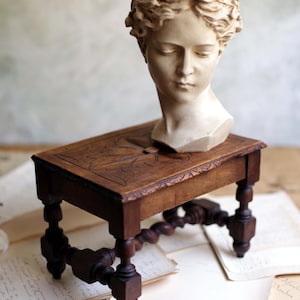 Peut inclure: Un buste en pierre beige représentant une personne couronnée de lauriers, posé sur un petit tabouret rectangulaire en bois brun foncé. Le tabouret présente quatre pieds tournés avec une entretoise torsadée et un motif de feuille sculpté.