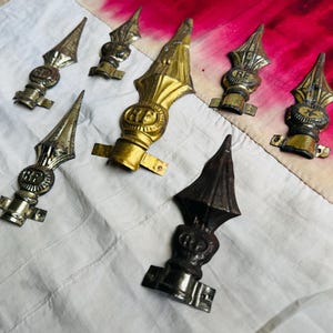 Puede incluir: Colección de terminales metálicos antiguos en tonos dorados y bronce. Cada terminal presenta un diseño de punta de lanza decorativa con detalles ornamentados y un emblema circular. Los terminales están dispuestos sobre una tela blanca y roja.