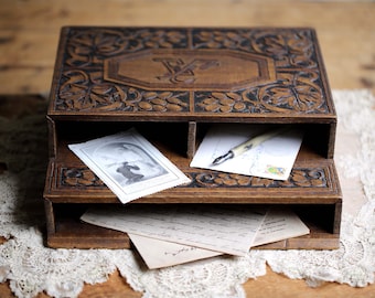May include: A vintage wooden letter holder with carved floral and geometric designs. The dark brown organizer has two shelves holding stationery, a pen, and a small picture. The top is decorated with the letter "V".