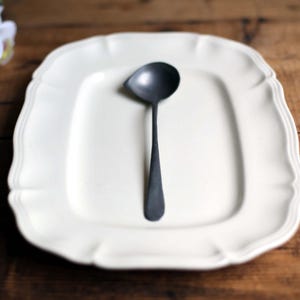 May include: A matte black ladle rests on a cream-colored, square plate with scalloped edges. The plate is centered on a wooden surface. The ladle's bowl is round, and the handle is long and slender.