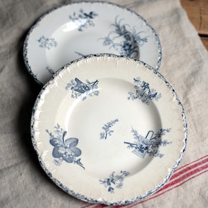 Peut inclure: Deux assiettes anciennes en céramique blanche avec des motifs floraux bleus. Les assiettes ont des bords festonnés et sont décorées de divers motifs floraux bleus, notamment des fleurs et des paniers. Les assiettes sont posées sur un tissu en lin.