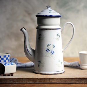 以下が含まれることがあります： 青い花柄と青い縁取りが施された白いエナメル製のコーヒーポット。ポットには蓋と注ぎ口が付いています。ポットは木製の表面の上に置かれています。