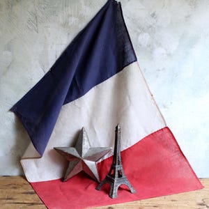 Può includere: Una bandiera francese con strisce verticali blu, bianche e rosse. Davanti alla bandiera si trovano una stella metallica a cinque punte con finitura testurizzata e una piccola replica dettagliata in metallo della Torre Eiffel grigio scuro.