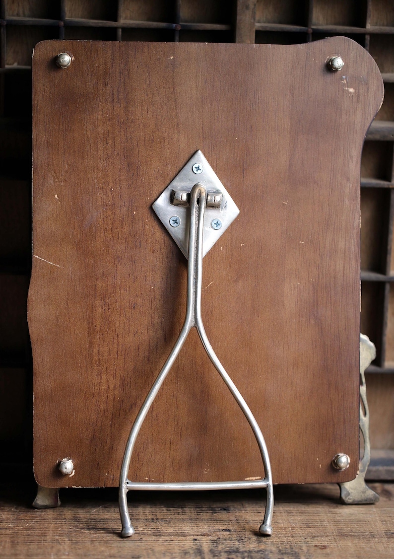 May include: A wooden picture frame with a silver metal stand. The stand is attached to the back of the frame with screws. The frame has a decorative design on the edges.
