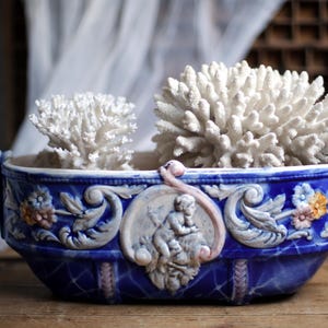 Può includere: Un vaso di ceramica blu con dettagli a rilievo ornati e manici, contenente due formazioni di corallo bianco. Il vaso presenta un rilievo centrale di cherubini e accenti floreali. L'estetica generale è vintage e decorativa.