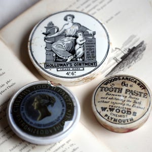 May include: Three antique tins with labels, resting on an open book. The largest tin, "Holloway's Ointment," features a black and white illustration. The other two are "Cherry Tooth Paste" and "Woods Areca Nut Tooth Paste."