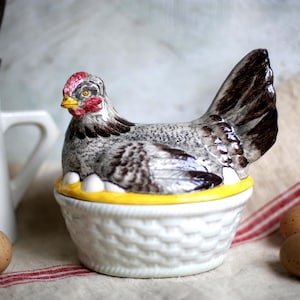 Op de afbeelding: Een keramische eierhouder in de vorm van een kip. De kip is grijs, wit en bruin, met een rode kam en gele snavel. Hij staat op een witte mand met een gele rand, met witte eieren. Bruine eieren liggen ernaast.