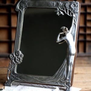 May include: A silver-toned, ornate mirror with a decorative frame featuring a stylized woman figure and floral motifs. The mirror is standing on a wooden surface.