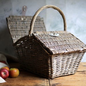 May include: A traditional wicker picnic basket, featuring a hinged lid and a curved handle. The basket is a natural brown hue, with a metal clasp. Several apples are placed on a wooden surface next to a white cloth.