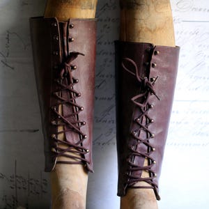 May include: Two antique, knee-high leather boots in a rich burgundy hue, laced up the front with matching cord. The boots are displayed on wooden shoe forms, with brass eyelets and a vintage aesthetic. The background features faded script.