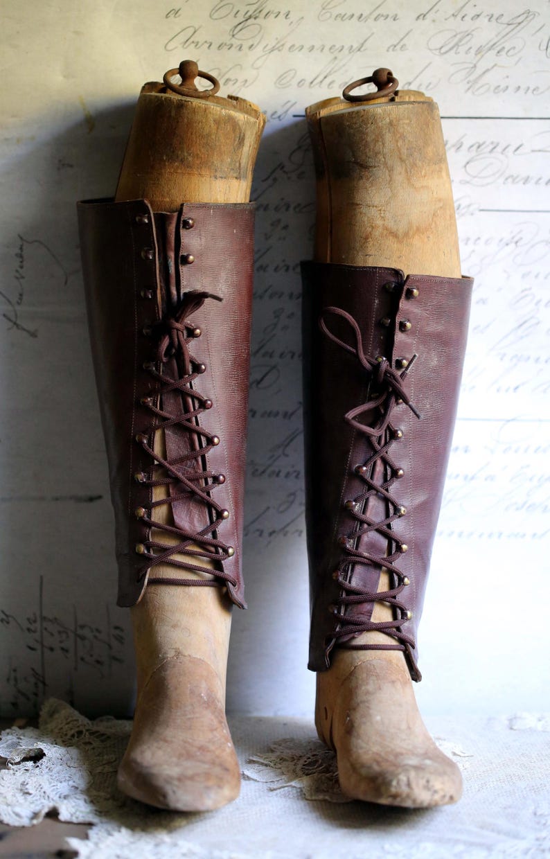 May include: Two antique, knee-high leather boots in a rich burgundy hue, laced up the front with matching cord. The boots are displayed on wooden shoe forms, with brass eyelets and a vintage aesthetic. The background features faded script.