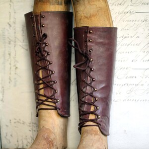 May include: Two vintage knee-high boots in a rich burgundy leather, laced up the front with brown cord. The boots are displayed on wooden shoe forms, showcasing their classic design. The background is a faded, antique-style print.