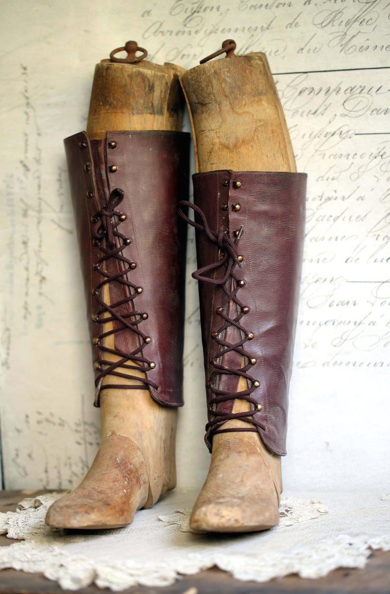 May include: Two vintage knee-high boots in a rich burgundy leather, laced up the front with brown cord. The boots are displayed on wooden shoe forms, showcasing their classic design. The background is a faded, antique-style print.