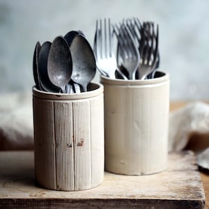 Könnte beinhalten: Zwei cremefarbene Keramik-Utensilienhalter gefüllt mit Besteck. Ein Halter enthält Löffel, der andere Gabeln. Die Halter sind zylindrisch mit vertikalen Rippendetails. Das Besteck hat ein Vintage-Aussehen mit Anlaufstellen.