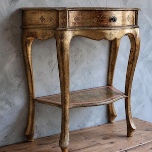 Puede incluir: Una mesa consola con acabado dorado, frente curvo, un cajón con un pequeño pomo oscuro y cuatro patas cabriolé. La mesa incluye un estante inferior con una superficie de patrón de diamantes y motivos florales decorativos en el marco.