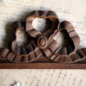 May include: A carved wooden decorative element with a ribbon design. The dark brown piece features a central oval and two looped sections, with a base. White daisies and handwritten paper are in the background.