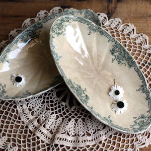 May include: Two oval-shaped ceramic dishes with a cream-colored base and a dark green decorative border featuring intricate scrollwork patterns. Each dish has a glossy finish and is accented with small white daisy flowers with black centers.