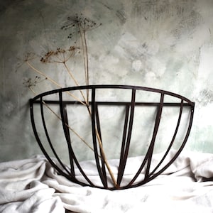 May include: A dark brown metal wall basket with a curved design, holding dried plant stems. The basket is set against a textured, muted green and gray background, resting on a white cloth.