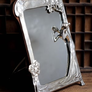 May include: A silver-toned, ornate mirror with a decorative frame featuring a stylized tree and a woman figure. The mirror has a beveled edge and stands on a wooden surface.