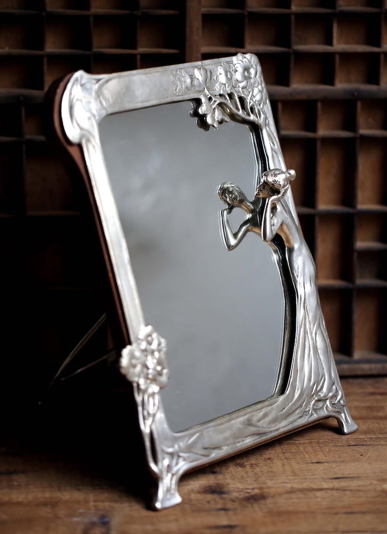 May include: A silver-toned, ornate mirror with a decorative frame featuring a stylized tree and a woman figure. The mirror has a beveled edge and stands on a wooden surface.