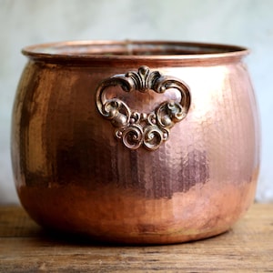 May include: A copper planter with a hammered finish and an ornate handle. The round planter has a wide opening and sits on a wooden surface. The handle has a bronze finish.