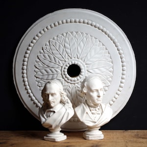May include: White plaster decorative ceiling medallion with a central floral design and two white plaster busts of men. The busts are on pedestals and the medallion is round. The background is black and the surface is wood.
