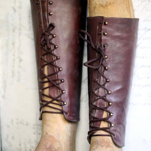 May include: Pair of antique, knee-high leather boots in a rich burgundy hue, laced up the front with dark brown cord. The boots are displayed on wooden shoe forms, showcasing their vintage design and craftsmanship. A classic piece.
