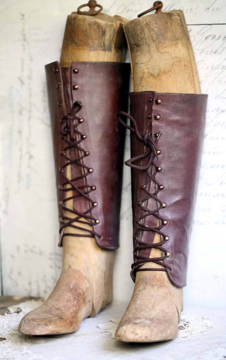 May include: Pair of antique, knee-high leather boots in a rich burgundy hue, laced up the front with dark brown cord. The boots are displayed on wooden shoe forms, showcasing their vintage design and craftsmanship. A classic piece.