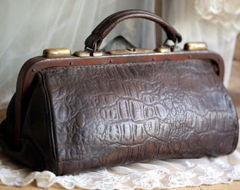 May include: A vintage brown leather doctor's bag with a textured crocodile pattern. The bag features a top handle, brass hardware, and a clasp closure. The bag is sitting on a white lace cloth.