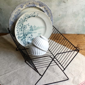 Peut inclure: Un égouttoir à vaisselle en métal noir présente deux assiettes anciennes avec des motifs floraux bleus et deux bols blancs côtelés. Les assiettes ont un bord festonné. L'égouttoir est posé sur un tissu en lin blanc.
