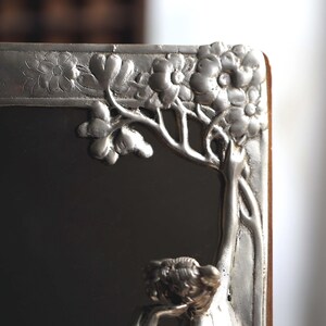May include: A silver-toned picture frame with an ornate floral design. The frame is made of metal and has a mirror inside. The frame is in the Art Nouveau style.
